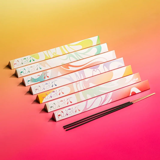 Six triangular prism boxes with colorful abstract swirl patterns are arranged diagonally on a yellow-to-pink gradient background, with three black incense sticks placed in front.