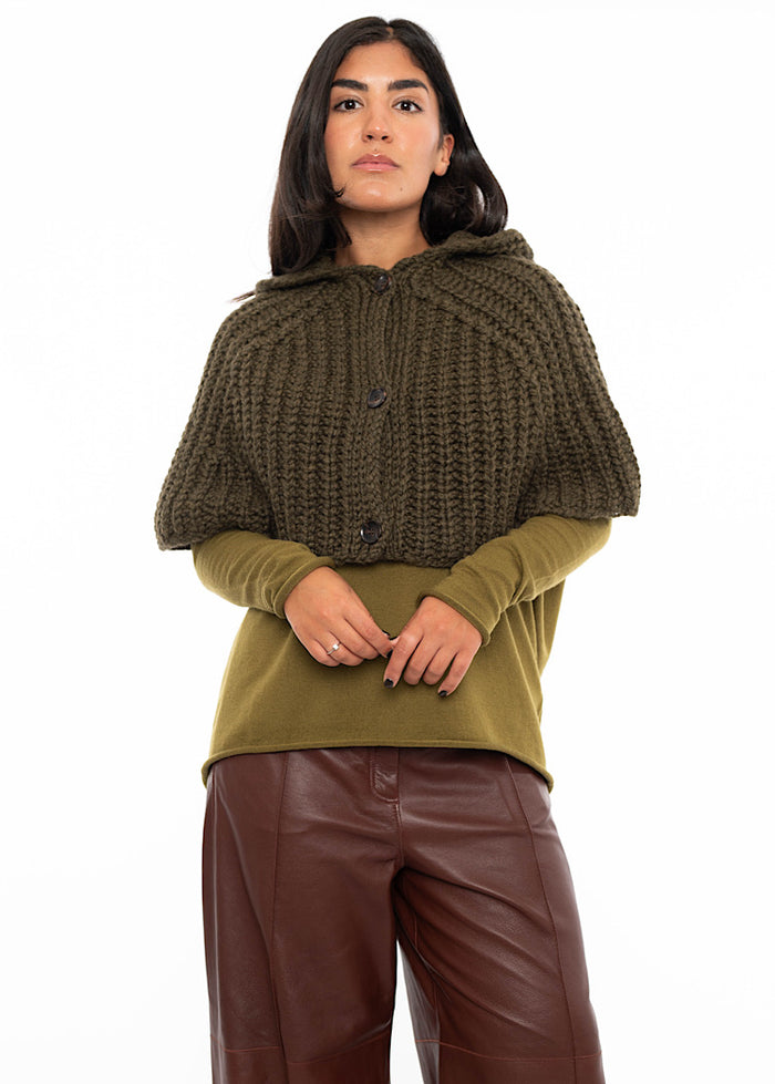 A model wearing an oversized knitted shawl with a hood in  dark green. Styled with a fitted khaki green pullover and burgundy leather trousers.