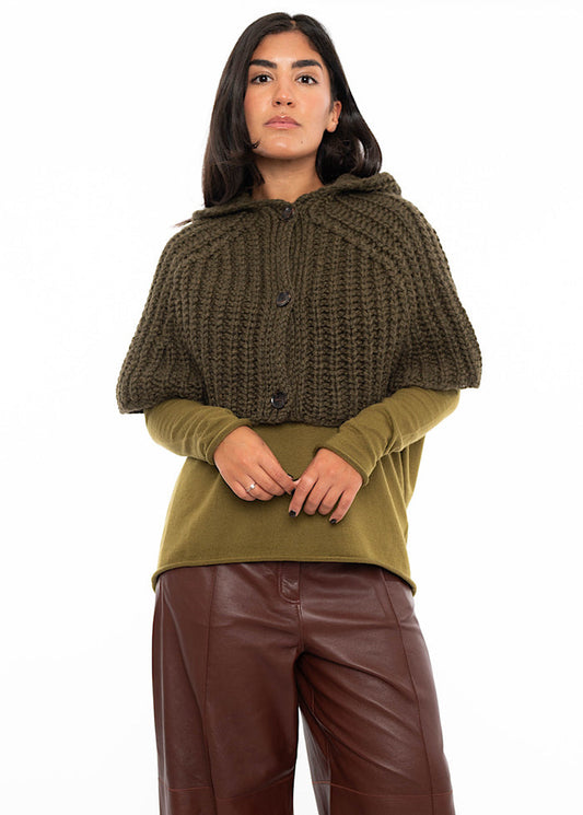 A model wearing an oversized knitted shawl with a hood in  dark green. Styled with a fitted khaki green pullover and burgundy leather trousers.