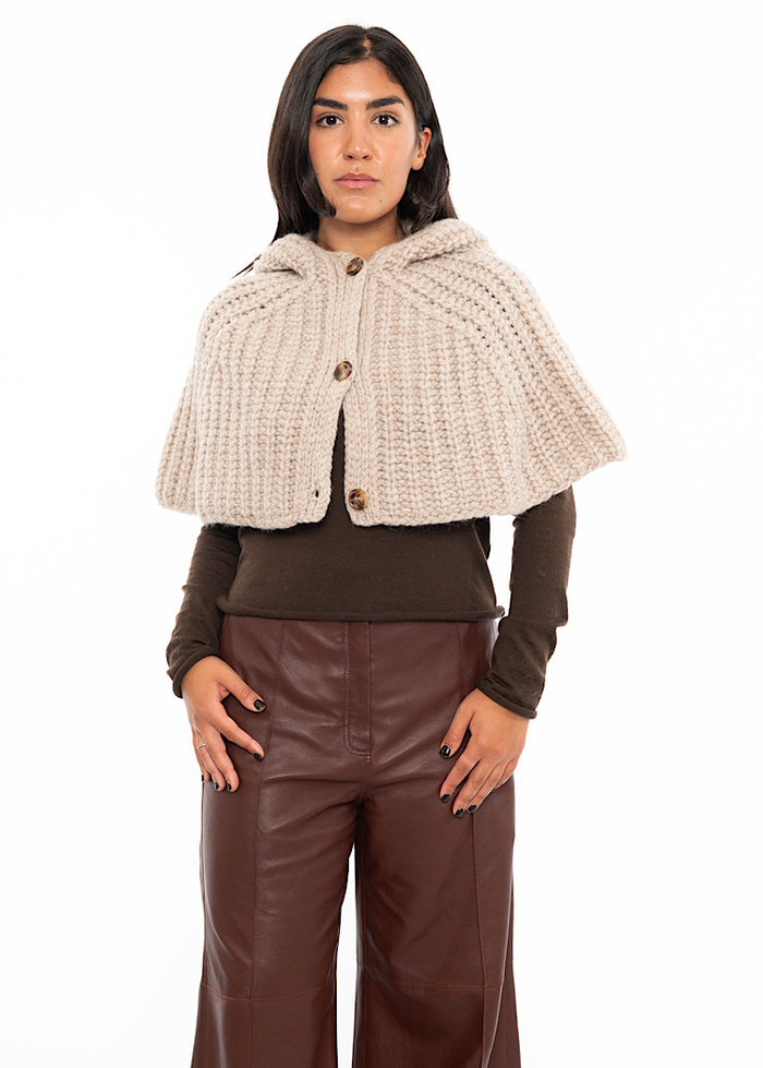 A model wearing an oversized knitted shawl with a hood in light beige. Styled with a fitted black pullover and Burgundy  leather jeans.
