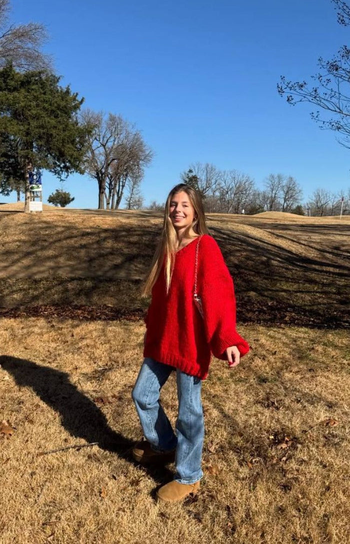 Red chunky knit sweater with a V-neck and oversized fit, showing long voluminous sleeves and ribbed cuffs outdoors