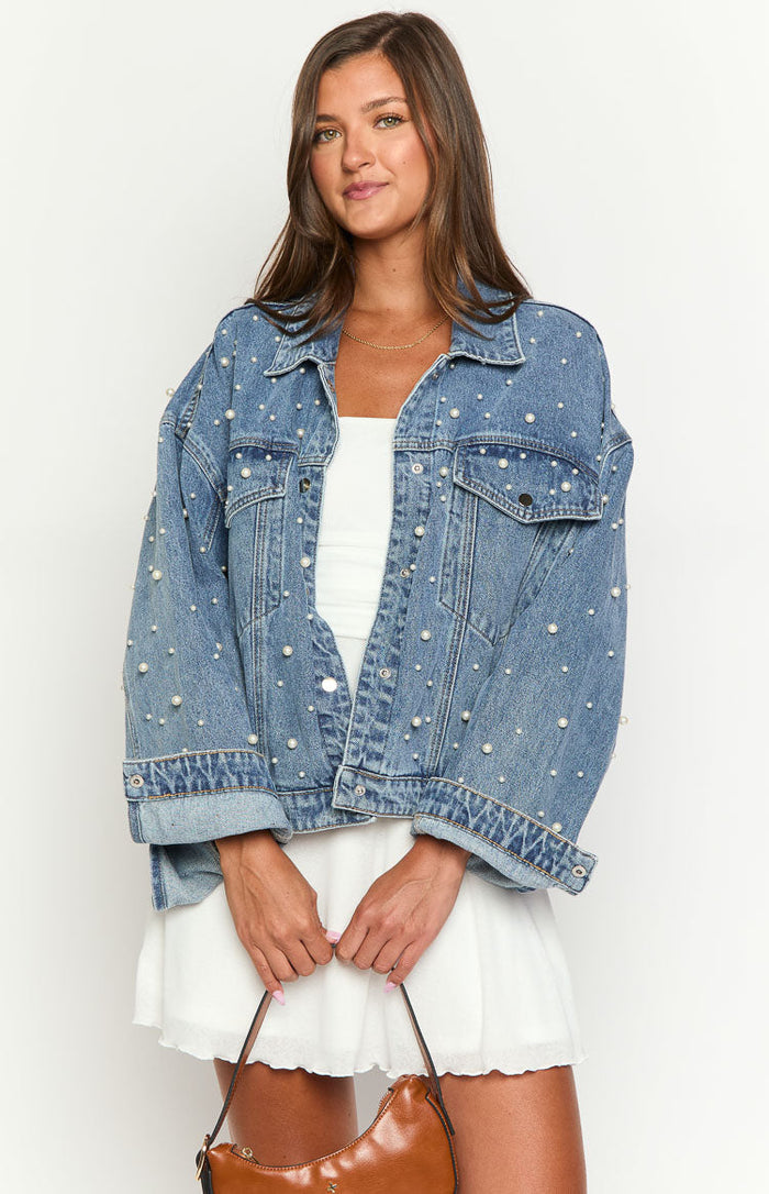 Front view of a mid wash denim jacket with an oversized fit, covered in small white pearl embellishments and button details