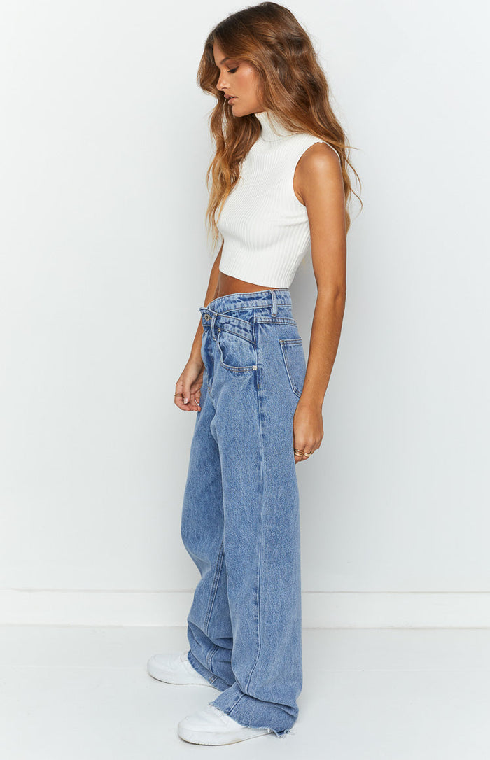 Side view of blue wide-leg jeans featuring an uneven crossover waist and a relaxed fit with a frayed hem
