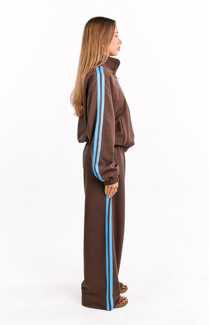 Side view of a chocolate brown relaxed fit jacket with blue sleeve stripes, high collar, ribbed cuffs, and side pockets