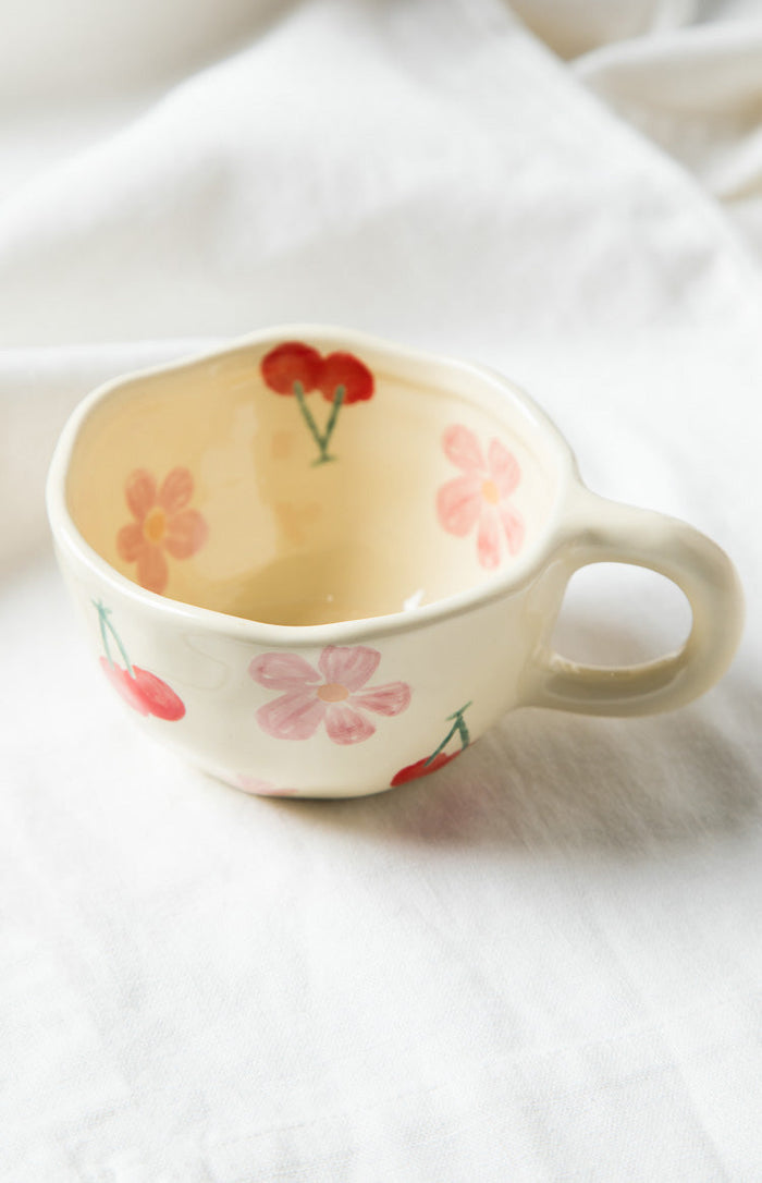 Cream ceramic mug with hand-painted pink flowers and red cherries, showing the handle and interior pattern