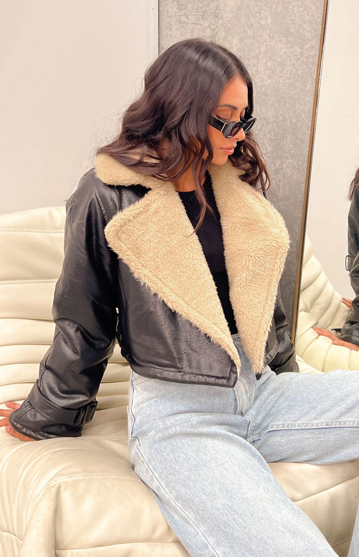 Side view of a black faux leather cropped jacket with a wide cream shearling collar and buckled cuffs