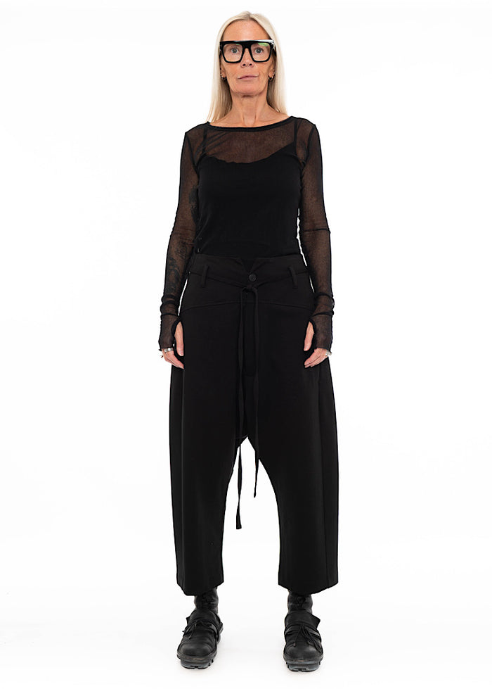 Model wearing black drop-crotch trousers with wide leg and drawstring detail, styled with mesh-sleeve top with combat boots.