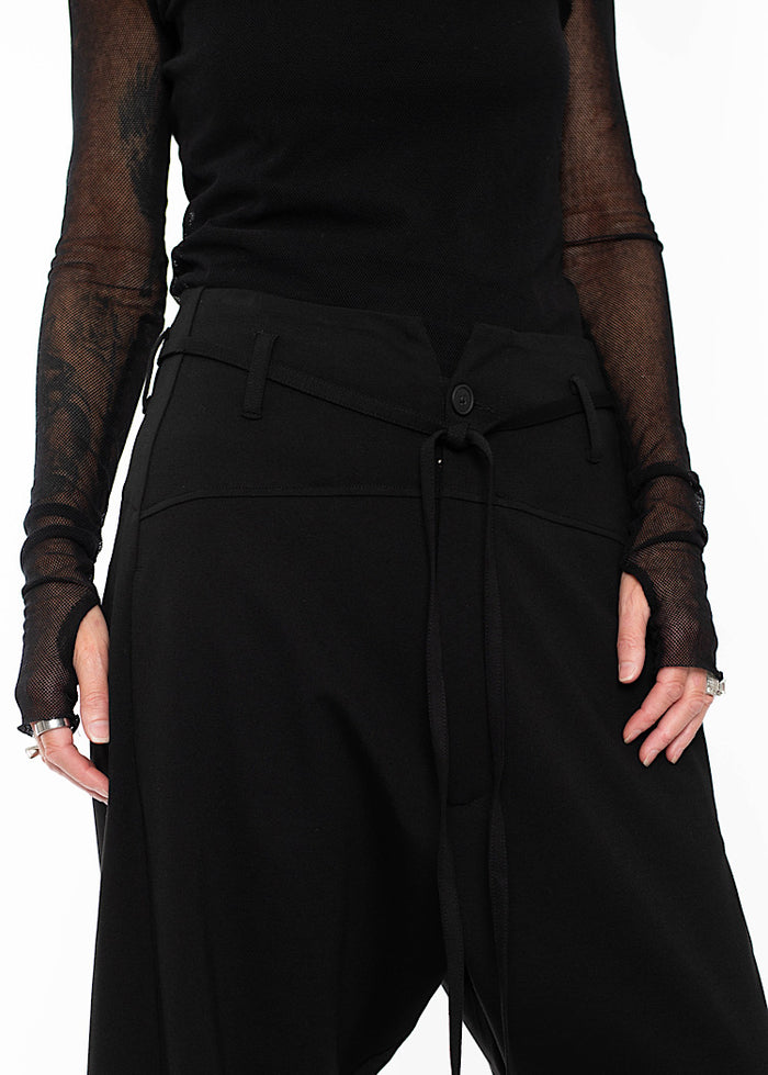 Close up of model wearing black drop-crotch trousers with wide leg and drawstring detail, styled with mesh-sleeve top and combat boots.