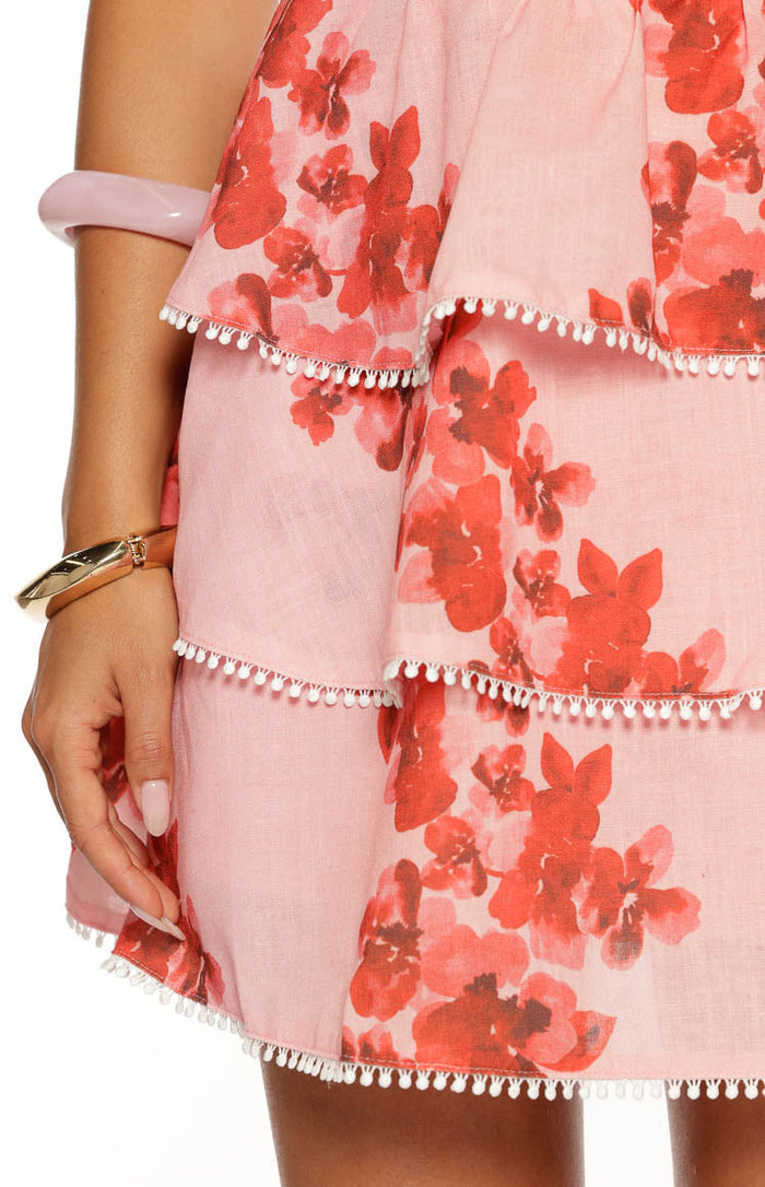 A close-up of the ruffled skirt of a mini-dress. The fabric is light pink and features a vibrant red and coral floral print. The skirt consists of multiple tiered ruffles each edged with a decorative white bobble or pom-pom trim.
