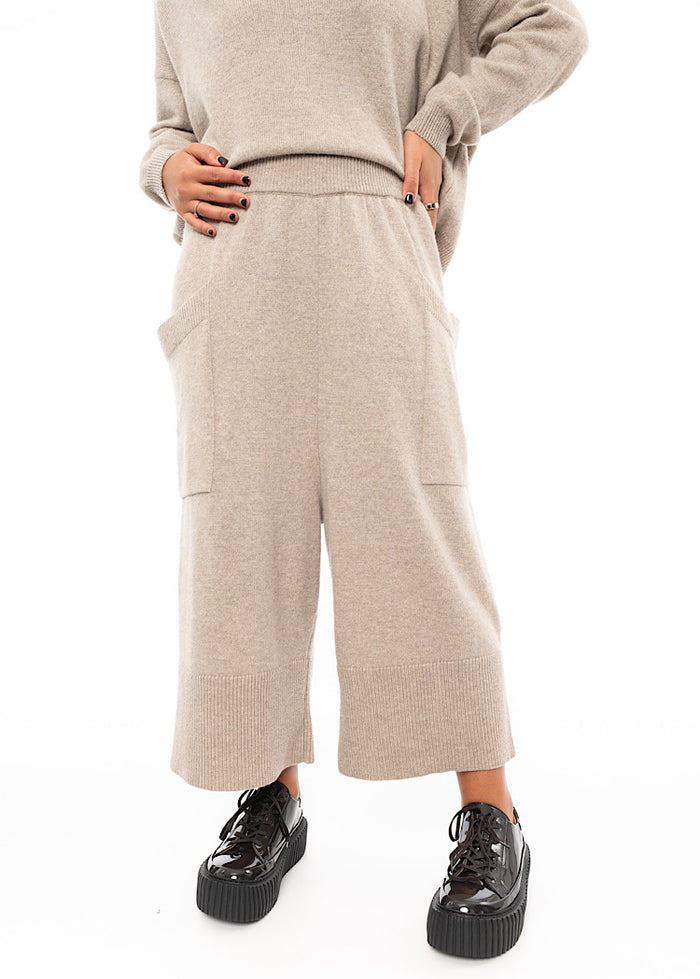 A Model wearing a light beige knitted Trouser. Paired with a matching pullover and Brown patent Shoes.