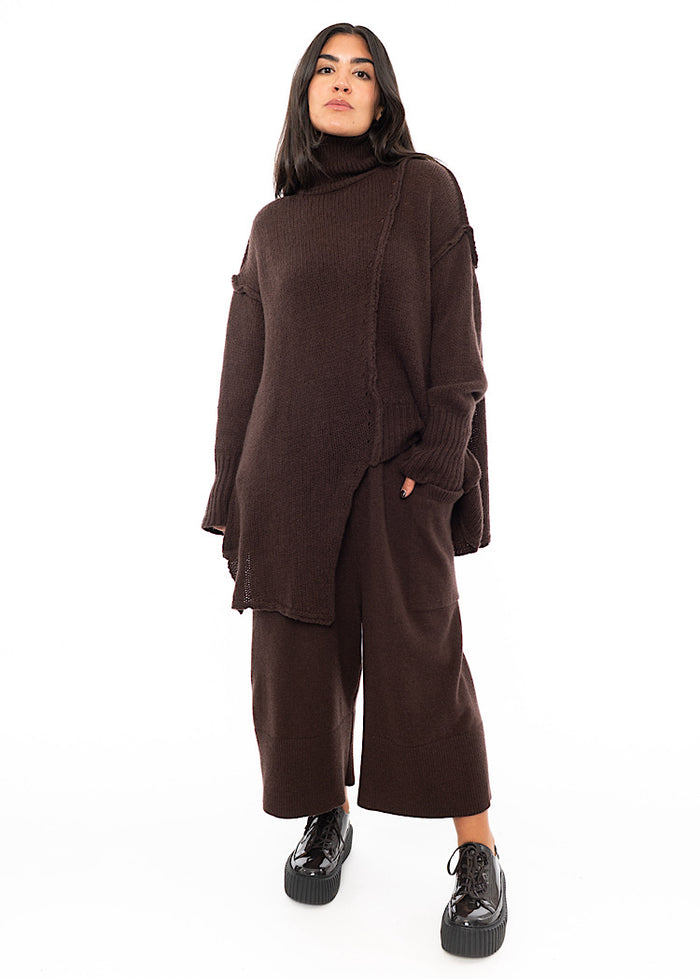 A Full frame photo of A Model wearing a Brown Oversized pullover featuring grungy pieces of fabric. Paired with a matching Trouser and Patent shoe.