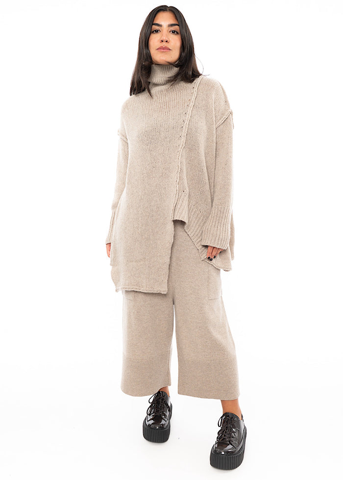 A full frame photo of A Model wearing a creme Knitted Pullover with  Distressed pieces throughout. Paired with a matching Trouser and patent Shoe.