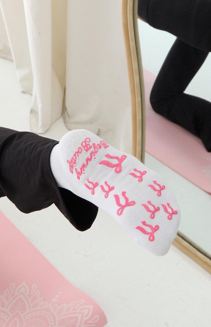 Bottom view of a white yoga sock with pink bow-shaped grip dots and Beginning Boutique branding on the sole