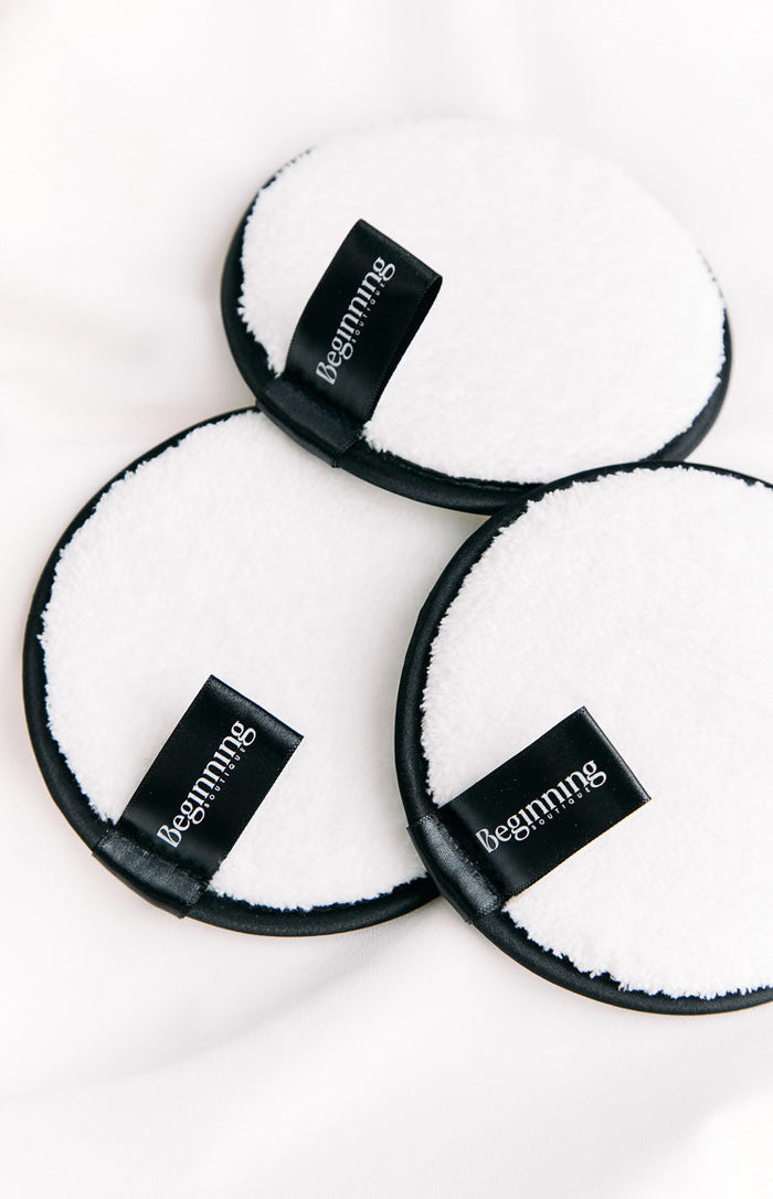 Close-up of three white circular reusable face wash pads with soft texture and black satin loop handles