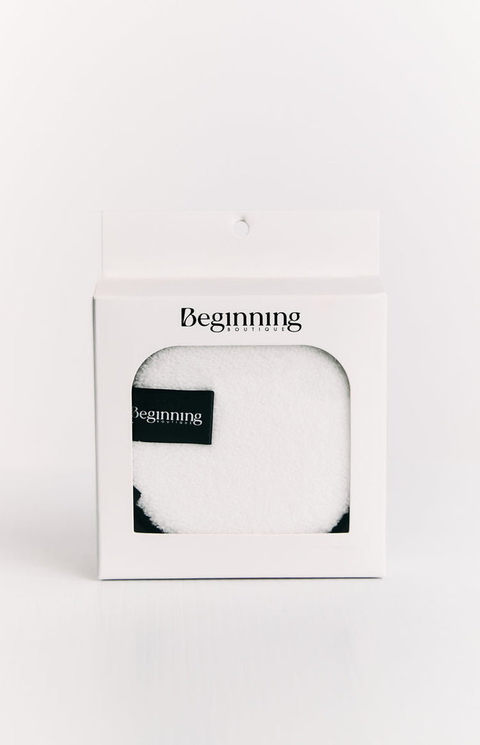 White fluffy reusable face wash pad with a black brand label, visible through the window of its white retail box