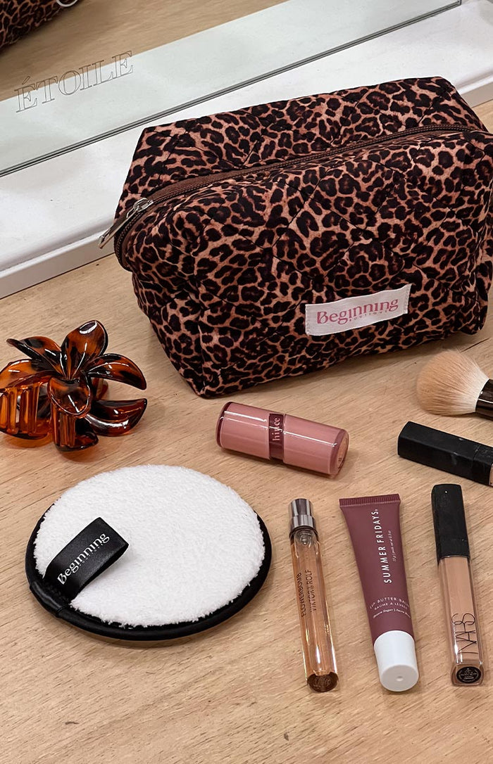 A round, white reusable makeup removing pad with a black border and a black 'Beginning' label, positioned near a quilted leopard print makeup bag and other cosmetics.